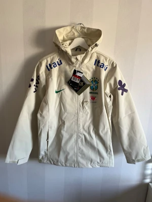 Nike Brasil vindjacka med huva - Stilren beige vindjacka från Nike med Brasil-logga och sponsormärken. Jackan har huva, dragkedja framtill och fickor på sidorna. Perfekt för dig som gillar sportig stil och vill visa ditt stöd för Brasilien. Materialet är lätt och skyddar mot vind.