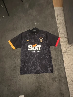 Galatasaray matchtröja Mertens #10 - Snygg svart Galatasaray matchtröja från Nike med Mertens och nummer 10 på ryggen. Tröjan har gul och röd detalj på ärmarna, klubbmärke och Nike-logga på bröstet samt Sixt sponsortryck. Tillverkad i lätt och ventilerande material, perfekt för fotboll eller träning.