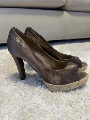 Bruna peep toe pumps från Replay - Snygga bruna pumps från Replay med peep toe och hög klack. Skorna har en platåsula med flätad detalj som ger en cool touch. Materialet är glansigt tyg och insidan är mjukt fodrad. Perfekta för dig som vill ha något extra till outfiten.