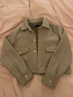 Beige kappa från Zara - Stilren croppad beige kappa från Zara med klassisk krage, silvriga knappar och två stora bröstfickor. Kappan passar perfekt nu inför våren och tidig höst. Knappt använd så därav väldigt bra skick.