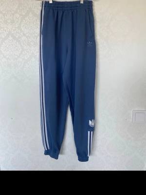Blå Adidas mjukisbyxor Primegreen - Snygga blå mjukisbyxor från Adidas med klassiska vita ränder längs sidorna och elastisk midja. Tillverkade i Primegreen-material och har Adidas logga på benet. Perfekta för chill dagar eller träning.