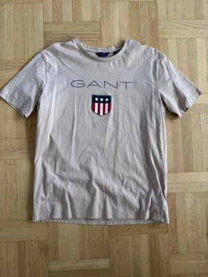 Beige GANT t-shirt med tryck - Snygg beige t-shirt från GANT med stort tryck framtill och logga med stjärnor och ränder. Kan gå ner i pris vid snabbt köp. Passar herr S också Xs