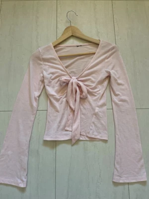 Ljusrosa långärmad topp med knut - Supersöt ljusrosa topp med långa ärmar och v-ringning. Toppen har ett knyt framtill som ger en extra detalj och är tillverkad i mjukt bomullsmaterial. Perfekt att styla med jeans eller kjol för en trendig look.