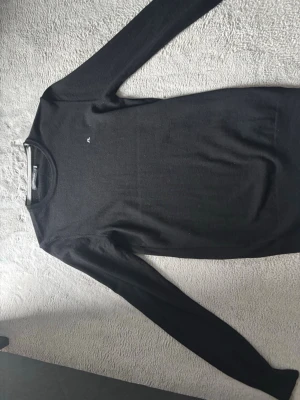 Svart stickad tröja från J.Lindeberg - Snygg svart stickad tröja från J.Lindeberg med rund halsringning och diskret logga broderad på bröstet. Tröjan har långa ärmar och är tillverkad i ett mjukt, tunt material som passar perfekt till lager på lager.