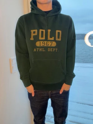 Mörkgrön hoodie från Polo Ralph Lauren - Mörkgrön hoodie från Polo Ralph Lauren med stor gul POLO 1967 ATHL. DEPT. print på bröstet. Klassisk känguruficka framtill och justerbar huva. Mjuk bomullsblandning och ribbade muddar vid ärmar och nederkant. Perfekt för en avslappnad och sportig stil.