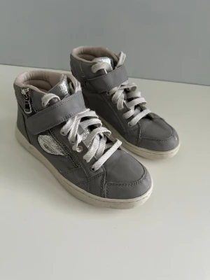 Gråa high top sneakers med dragkedja, storlek 35 - Snygga grå sneakers med högt skaft, vita snören och kardborreband. Skorna har silverfärgade detaljer, dragkedja på sidan och en glansig metallicdetalj. Yttermaterialet är troligtvis syntet eller läderimitation. Perfekta för en trendig streetstyle-look.