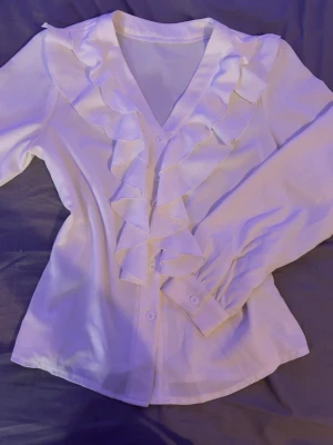 Vit blus med volanger och v-ringning - Stilren vit blus med långa ärmar och breda manschetter. Blusen har en djup v-ringning och dekorativa volanger längs knäppningen framtill. Tillverkad i ett lätt, mjukt tyg som ger ett elegant intryck. Perfekt för att lyfta vilken outfit som helst.