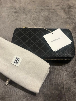 Svart smyckesväska By Malene Birger - Snygg svart smyckesväska från By Malene Birger med guldfärgad dragkedja och mönstrad yta. Insidan har flera fack och ringhållare i svart sammet. Perfekt för att organisera smycken. Medföljer tygpåse med logga. Helt ny och oanvänd, kunde inte göra reatur.