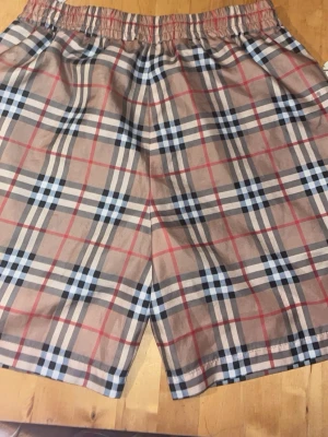 Burberry shorts  - Säljer ett par klassiska rutiga shorts från Burberry i beige, svart, vit och röd. Shortsen har elastisk midja och är tillverkade i bomull med meshfoder. Perfekta för sommaren och har den ikoniska Burberry-looken.