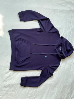 Mörkblå hoodie från Polo Ralph Lauren - Snygg mörkblå hoodie från Polo Ralph Lauren med klassisk huva och dragsnören. Har en känguruficka framtill och den ikoniska ljusblå Polo-loggan broderad på bröstet. Tillverkad i mjukt bomullsmaterial som är skönt att bära.