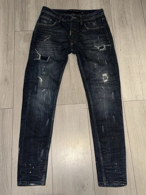 Mörkblå distressed jeans  - Säljer ett par mörkblå jeans från Alessandro zavetti med slim fit passform. Jeansen har slitningar, färgstänk och patchade detaljer för en riktigt edgy look. Klassisk femficksmodell med dragkedjegylf och cool streetkänsla. Perfekta för dig som gillar statement-plagg.