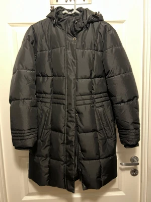 Svart lång pufferjacka från Take Off - Säljer en svart lång pufferjacka från Take Off med huva och dragkedja framtill. Jackan har två fickor, quiltad design och justerbar huva. Perfekt för kalla vinterdagar och riktigt skön att bära. Materialet är glansigt och ger en modern look. Den har ett hål i vänstra ärmen som går att sy som ny.