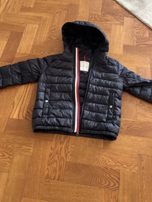 Svart dunjacka från Moncler med huva - Snygg svart dunjacka från Moncler med huva och quiltad design. Jackan har två framfickor med tryckknappar och dragkedja med rödvit detalj. Moncler-logga på ärmen ger en exklusiv känsla. Perfekt för alla dagar under året