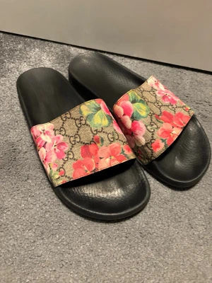 Gucci sandaler med blommönster - Säljer ett par svarta sandaler från Gucci med bred rem i canvas med klassiskt GG-mönster och färgglada blommor i rosa, rött och grönt. Snygg och trendig design som sticker ut. Sulan är i svart gummi och remmen har en lyxig känsla. Remmen i läder. Sparsamt använda. Motsvarande pris idag begagnat mycket högre. Äkthetsbedömd och köpt i Sverige 
