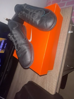 Svarta Nike fotbollsskor med blå detalj - Säljer ett par svarta fotbollsskor från Nike med blå swoosh på sidan. Skorna har snörning och är tillverkade i syntetmaterial med en modern, sportig design. Perfekta för dig som vill ha stilrena och funktionella skor på planen.