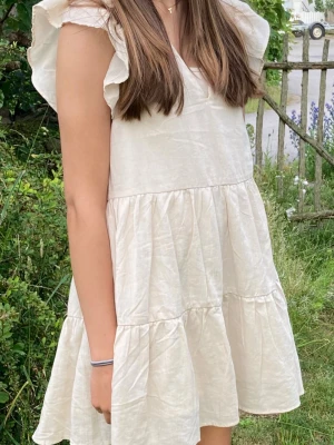 Beige kortklänning i linne - Söt beige kortklänning i luftigt linne med volanger och avslappnad passform. Klänningen har ärmlös design och en v ringning ger en somrig känsla. Perfekt för varma dagar och enkel att styla med accessoarer. Från hm endast använd en gång