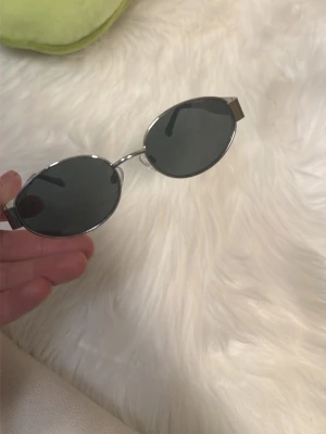 Ovala solglasögon med silverbåge - Snygga ovala solglasögon med mörka glas och tunn silverfärgad metallbåge. Glasögonen har en minimalistisk och trendig design som passar perfekt till sommaren. Inget synligt märke på bågen.