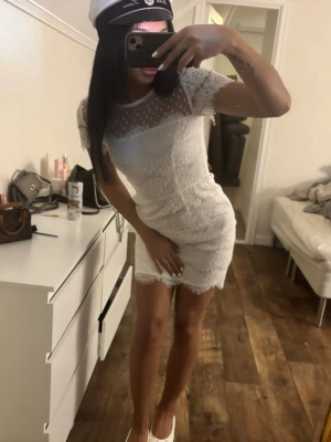 Ida sjöstet student klänning  - Kort vit spetsklänning med figurnära passform och korta ärmar. Överdelen har transparent mesh med prickigt mönster och rund halsringning. Perfekt till student eller sommarens tillfällen. Klänningen har fina spetsdetaljer och är riktigt söt och trendig.