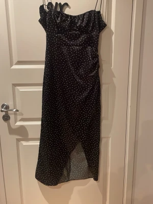 Svart prickig långklänning med slits från Zara - Svart långklänning med vita prickar och tunna axelband. Klänningen har en lätt glansig finish, omlottdetalj framtill och en liten slits. Öppen rygg med snörning. Snygg drapering vid bysten och smickrande passform. Perfekt för dig som gillar en klassisk men trendig look.