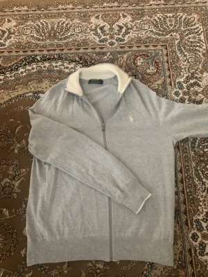 ziptröja från Polo Ralph Lauren - Snygg ljusgrå ziptröja från Polo Ralph Lauren med vit ribbad krage och diskret broderad logga på bröstet. Tröjan har hel dragkedja, långa ärmar och ribbade muddar. Perfekt för en avslappnad men stilren look.