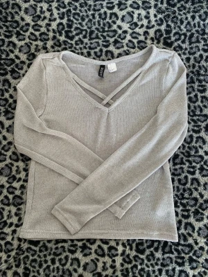 Ljusgrå stickad tröja Divided H&M - Snygg ljusgrå stickad tröja från Divided H&M med v-ringning och dekorativ banddetalj framtill. Tröjan har lång ärm och är tillverkad i ett tunt, mjukt material som passar perfekt till lager på lager. Enkel och stilren design som är lätt att matcha.
