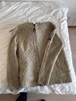 Beige stickad kofta med dragkedja - Säljer en beige stickad kofta med dragkedja och snyggt kabelstickat mönster. Koftan har ribbade muddar, fickor och en liten läderdetalj med logga vid halsen. Perfekt för lager på lager och riktigt nice till våren