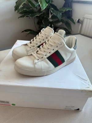 Gucci vita sneakers med gröna/röda ränder - Säljer ett par klassiska vita sneakers från Gucci med gröna och röda sidoremsor samt grön detalj vid hälen. Skorna är i läder och har en låg profil med snörning. Perfekta för dig som vill ha en stilren och ikonisk look.