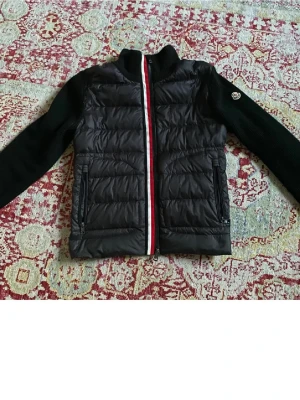 Svart cardiganjacka från Moncler - Snygg svart cardiganjacka från Moncler med quiltad front och stickade ärmar. Dragkedja framtill med röd, vit och blå rand. Två fickor med dragkedja och Moncler-logga på ärmen. Perfekt för lager på lager och streetstyle. Finns en liten defekt på högra bröstet det är lagat men den finns där men inget man märker vid användning.
