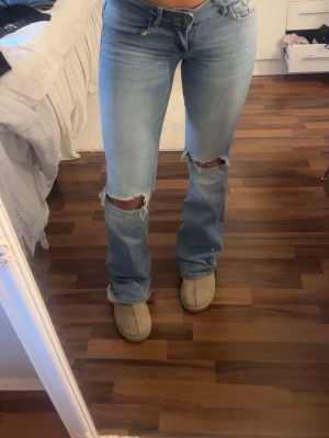 Ljusblå bootcut med hål i  - Säljer ett par ljusblå bootcut jeans med slitningar över knäna och snygga bakfickor med lock. Jeansen har en klassisk femficksmodell och är tillverkade i denim med stretch för skön passform. Perfekta för en avslappnad och trendig look.