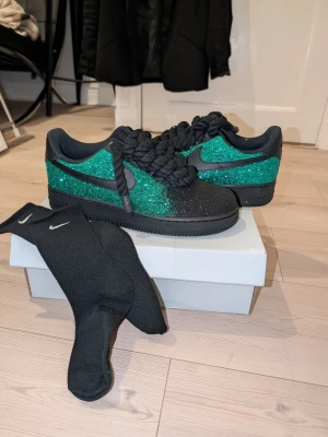 Nike Air Force 1 glitter oanvända - Unika Nike Air Force 1 sneakers med svart och grön glitterfinish. Skorna har tjocka svarta snören och klassisk låg siluett. Swoosh-loggan på sidan och svart sula ger en clean look. Materialet är textil och syntet med glittrig yta. Perfekta för dig som vill sticka ut. Aldrig använt 