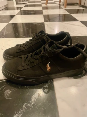 Svarta Polo Ralph Lauren sneakers - Snygga svarta sneakers från Polo Ralph Lauren med klassisk låg profil. Skorna har svart canvasmaterial, matchande svarta skosnören och den ikoniska broderade Polo-loggan i vitt och guld på sidan. Yttersulan är brun med Polo-tryck undertill.