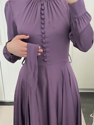 Lila långklänning med knappar - Elegant lila långklänning med höga knappar framtill, långärmad design och matchande bälte i midjan. Klänningen har rynkad halsringning och breda manschetter med klädda knappar. Materialet har en mjuk, lätt glansig finish. Kolla i min profil för andra klänningar också!! finns även i mintgrön