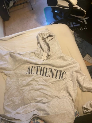 Grå hoodie med AUTHENTIC-tryck - Snygg ljusgrå hoodie med stor huva och texten AUTHENTIC tryckt i svart på bröstet. Hoodien har korta ärmar och ribbad nederkant. Perfekt för en avslappnad streetwear-look. Materialet känns mjukt och bekvämt, troligen bomullsblandning.