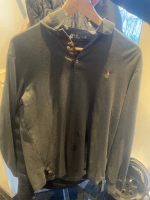 Mörkgrå långärmad tröja Polo Ralph Lauren - Snygg mörkgrå långärmad tröja från Polo Ralph Lauren i custom slim fit. Tröjan har klassisk krage med knappar och den ikoniska broderade loggan på bröstet. Perfekt till både jeans och chinos för en clean look.