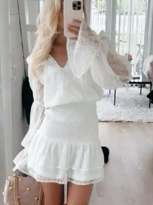 Vit spetsklänning med volang - Supersöt vit kortklänning från in the style med transparenta spetsärmar, v-ringning och volangdetaljer. Klänningen har smockad midja och är perfekt för studenten eller bara till sommaren💕