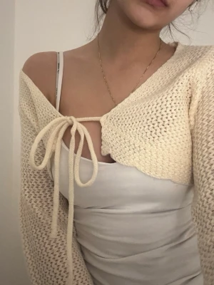 Beige stickad croppad kofta med knyt - Superfin beige stickad kofta med croppad passform och långa ärmar. Koftan har en snygg knytning framtill och är perfekt att slänga över ett linne eller klänning. Mjuk och mysig, passar perfekt för lager-på-lager-stil.