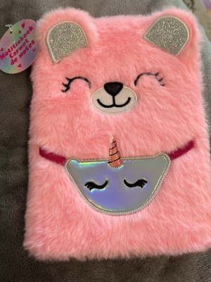 Anteckningsbok med rosa fluff och björn - Säljer en supergullig anteckningsbok med mjukt rosa fluffigt omslag, dekorerad med ett björnmotiv och glittriga öron. Framsidan har även en holografisk enhörningsdetalj. Perfekt för att skriva ner tankar, idéer eller skisser. Linjerade sidor inuti.
