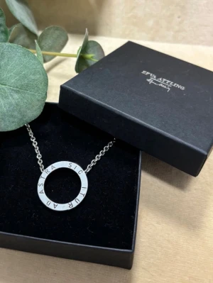 Silverhalsband med cirkelberlock - Efva attling halsband. Ett nyskick halsband i silver med en rund cirkelberlock där texten 'AD ASTRA SIC ITUR' är ingraverad runt hela ringen. Kedjan är tunn och elegant, perfekt för dig som gillar minimalistiska smycken med en personlig touch. Original pris 2500 kr, nu 1600.