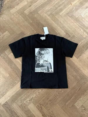 Maison Margiela svart t-shirt - Svart t-shirt från Maison Margiela med en stor grafisk print framtill i svartvitt. Klassisk passform med korta ärmar och rund halsringning. Trycket har siffror och texten 'Maison Margiela Paris'. Perfekt för dig som gillar statement-plagg. Passar någon runt 178-184