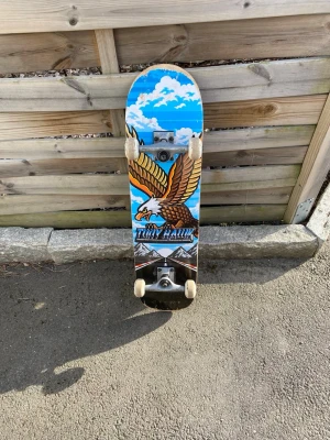 Tony Hawk skateboard med örn - Säljer en skateboard från Tony Hawk med en färgglad design av en örn mot blå himmel och moln. Brädan har vita hjul och svart grepptejp på ovansidan. Perfekt för dig som vill cruisa eller öva tricks i skateparken.