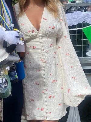 Adoore klänning  - Söt vit kortklänning med små röda blommor, v-ringning och knappar framtill. Klänningen har långa, vida ärmar och omlottdetalj vid bysten. Perfekt för sommarens alla tillfällen.