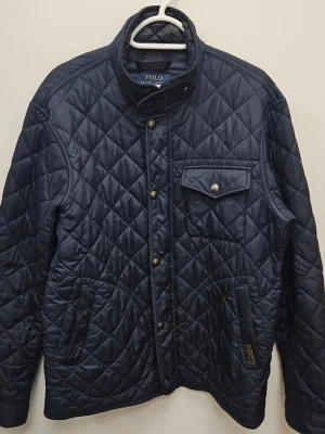 Marinblå quiltad jacka Polo Ralph Lauren - Snygg marinblå quiltad jacka från Polo Ralph Lauren med tryckknappar, ståkrage och fickor fram. Jackan har ett rutmönster och en bröstficka med lock. Perfekt för dig som gillar klassisk och stilren design.