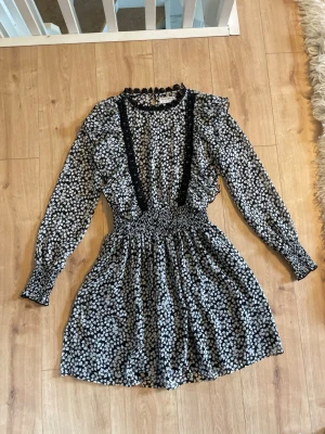 Mönstrig kortklänning från Holly & Whyte - Supersöt kortklänning från Holly & Whyte med svartvitt mönster, långa ärmar och smock i midjan. Klänningen har volangdetaljer på axlarna, spets längs bröstet och en liten volangkrage. 