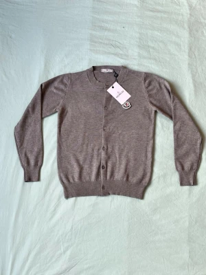 Beige kofta från Moncler - Snygg beige kofta från Moncler med rund halsringning och knäppning framtill. Koftan har Moncler-logga på bröstet och ribbade muddar vid ärmslut och nederkant. Tillverkad i mjukt material som känns lyxigt och stilrent.