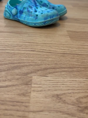 Blå tie-dye tofflor från H&M - Säljer ett par blå tofflor från H&M med tie-dye mönster i olika blå nyanser. Tofflorna har klassisk crocs-design med hål på ovansidan och justerbar hälrem. Tillverkade i lätt plastmaterial som är enkla att rengöra.