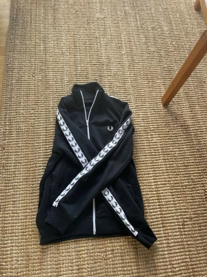 Svart Fred Perry ziptröja - Säljer en svart ziptröja från Fred Perry med vita detaljer. Tröjan har vita dragkedjor, ståkrage och vita ränder med Fred Perry-loggor längs ärmarna. Klassisk och sportig design, perfekt för en avslappnad streetwear-look. Knappt använd 