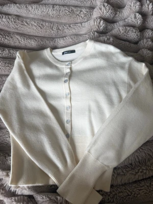 Begie kofta från Gina  - Snygg begie kofta från gina tricot med rund halsringning och knappar framtill. Tröjan har långa ärmar och bred ribbad kant nedtill för en modern look. Perfekt att slänga över en topp eller klänning för extra stilpoäng.