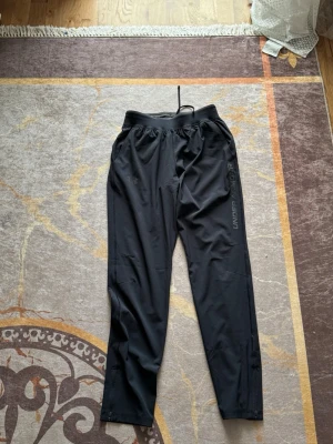 Svarta träningsbyxor Under Armour - Säljer ett par svarta träningsbyxor från Under Armour med resår i midjan och diskret logga längs benet. Byxorna är tillverkade i ett lätt och stretchigt material som passar perfekt för gym, löpning eller annan träning.