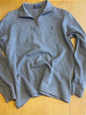 Grå quarter zip från Polo Ralph Lauren - Snygg grå quarterzip tröja från Polo Ralph Lauren med quarter zip och brunt läderdetalj på dragkedjan. Klassisk broderad logga på bröstet och ribbade muddar. Perfekt till jeans eller chinos för en clean look. 70 cm längd, 58 cm axel till axel