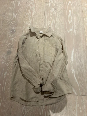 Beige linneskjorta från Lindex - Snygg beige skjorta från Lindex i storlek 152. Tillverkad i linne med klassisk krage, bröstficka och knäppning framtill. Perfekt för en avslappnad men ändå stilren look. Långärmad och i en neutral färg som passar till det mesta.